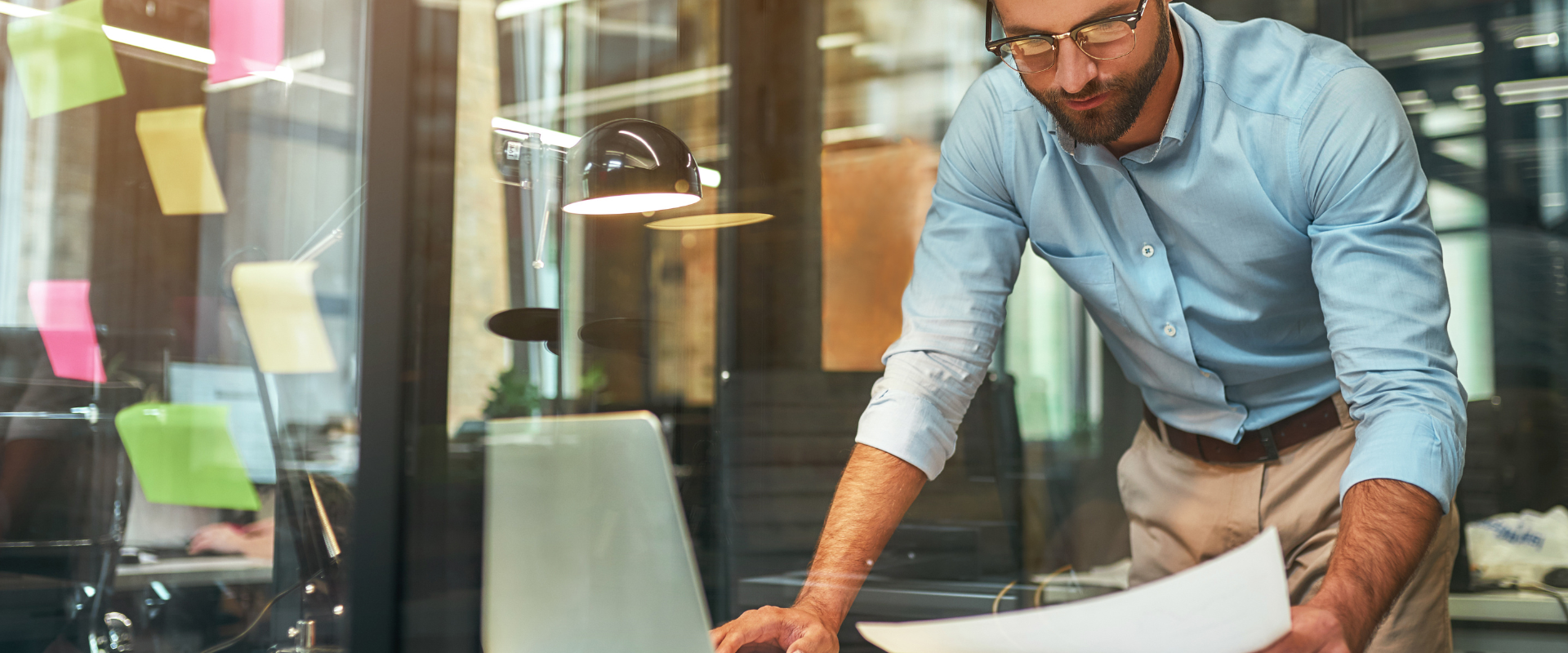 Junger bärtiger Geschäftsmann mit Brille und formeller Kleidung, der im modernen Büro stehend Dokumente prüft.