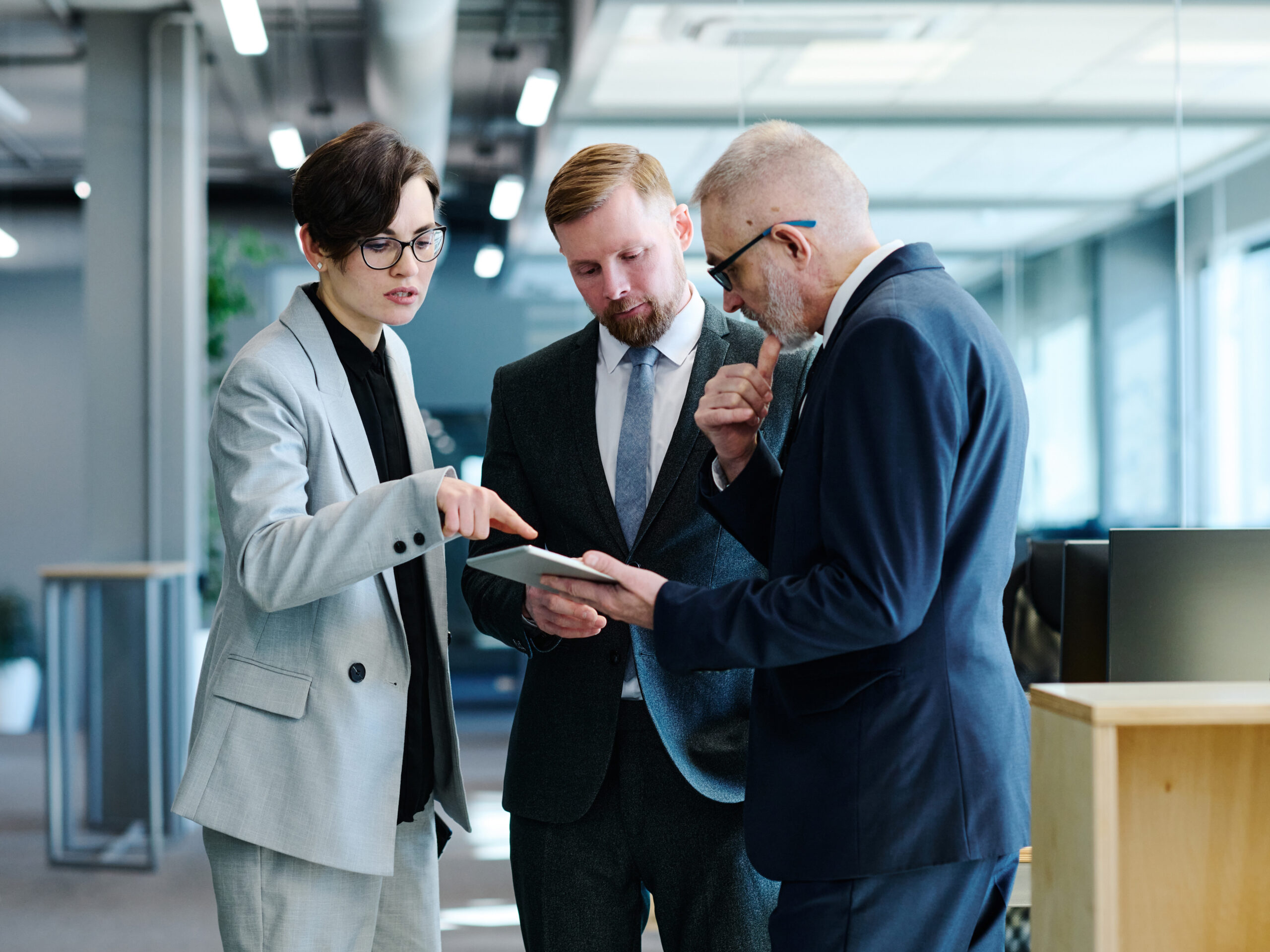 Eine Frau und zwei Männer haben Anzügen an und stehen in einem Büro. Sie schauen sich etwas auf einem Tablet an. 