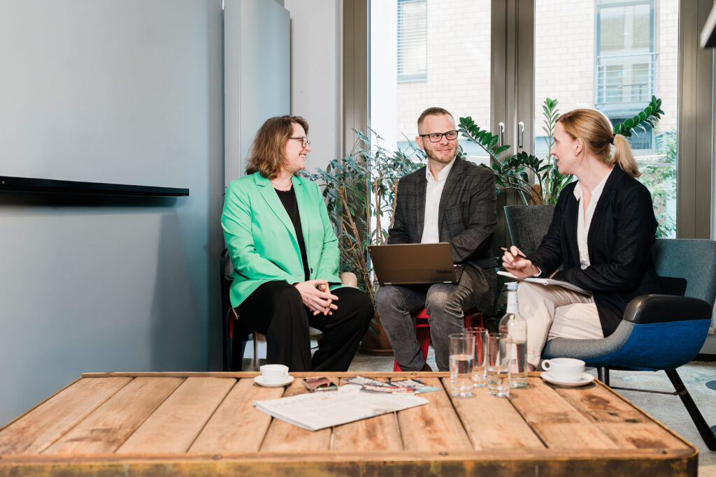 Zwei Frauen und ein Mann sitzen auf Stühlen in einem Bürogebäude und unterhalten sich. 